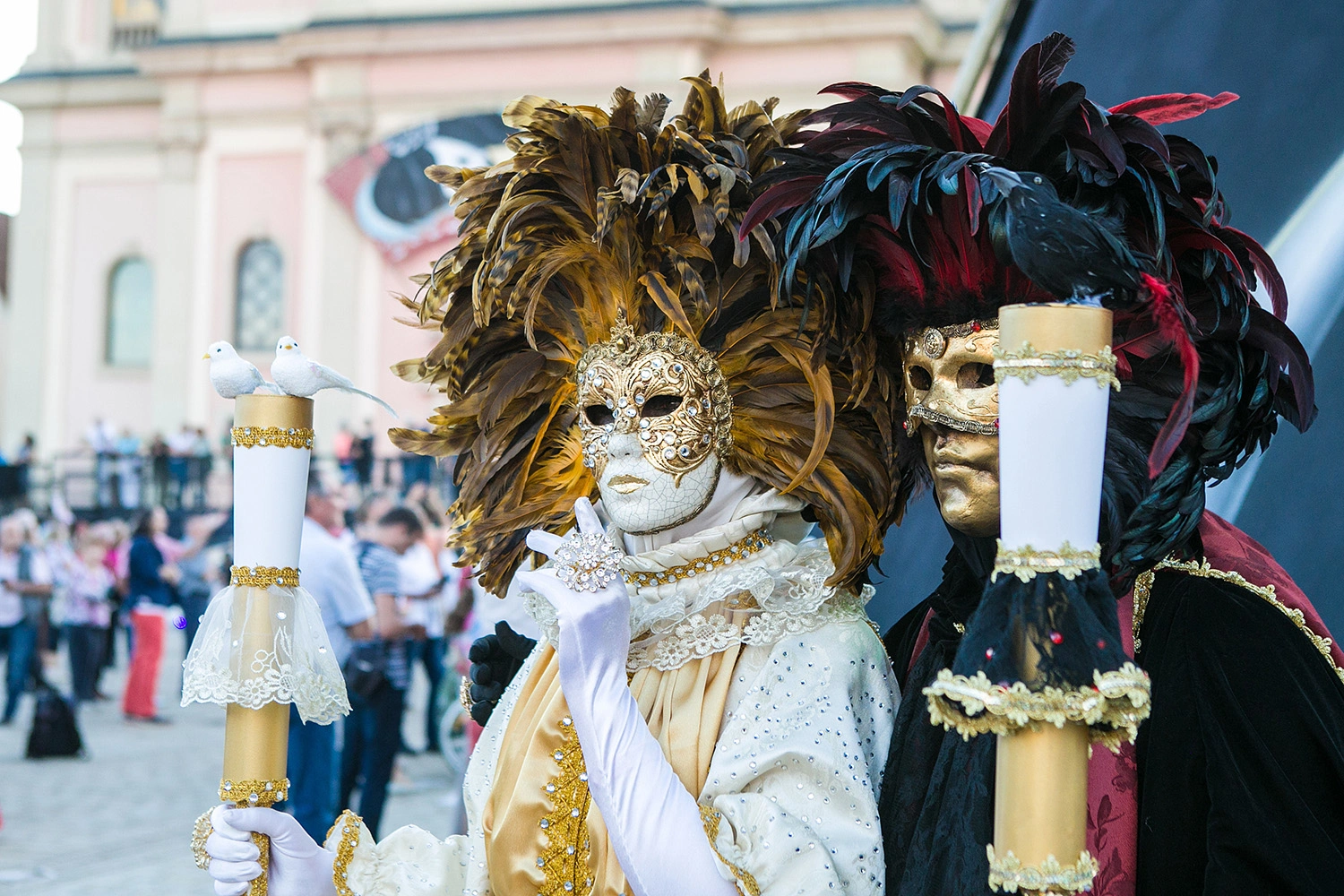 Zwei Personen in aufwendigen Masken und Federkostümen posieren beim venezianischen Karneval auf einem Platz vor historischer Kulisse.