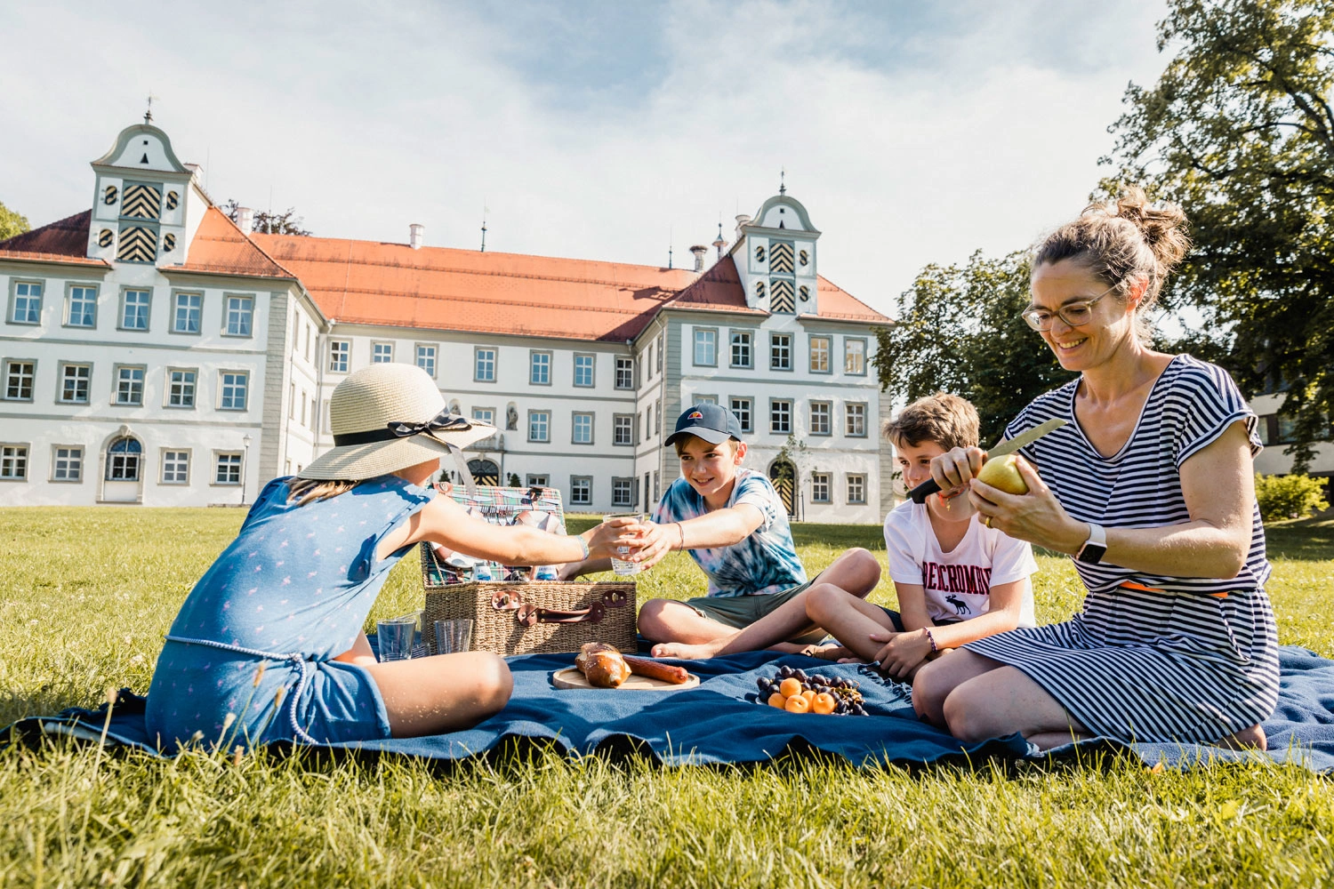 Eine Frau sitzt mit ihren Kindern auf einer Decke in einem Park vor einem Schloss und picknickt.