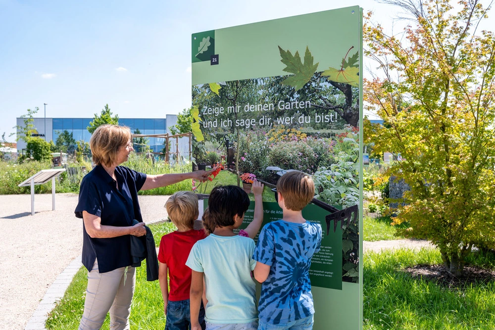 A woman stands with a group of children by a sign in a garden and explains its contents.