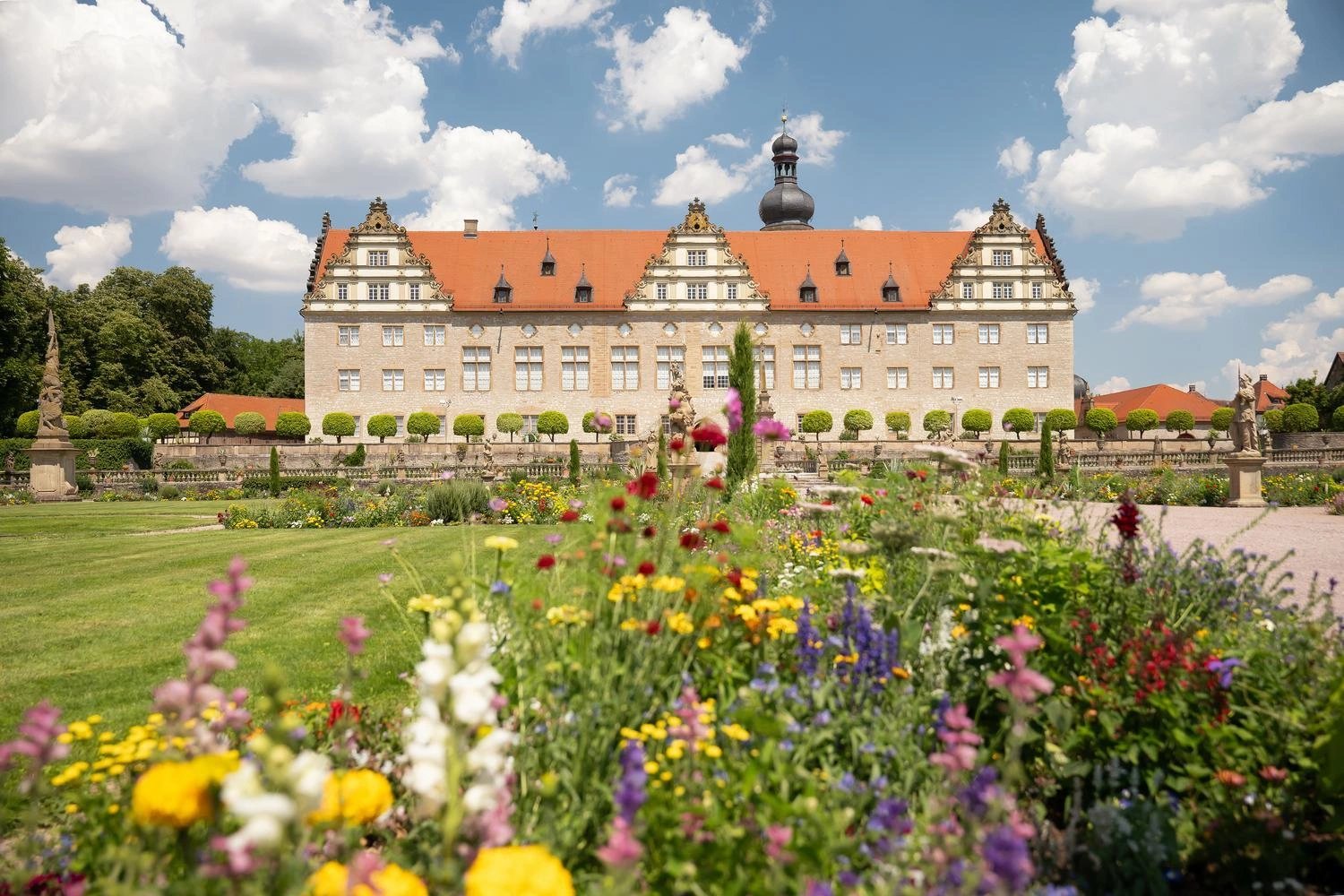 Baroque castle with red roof and towers, surrounded by a well-kept garden with colorful flowers in the foreground under a blue sky with white clouds.
