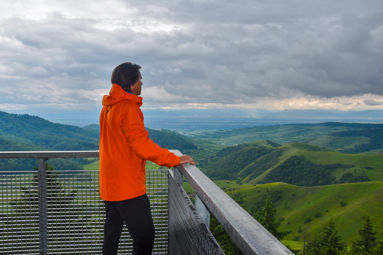Ein Mann in orangener Jacke steht auf einem Aussichtsturm am Geländer und schaut in die Ferne.