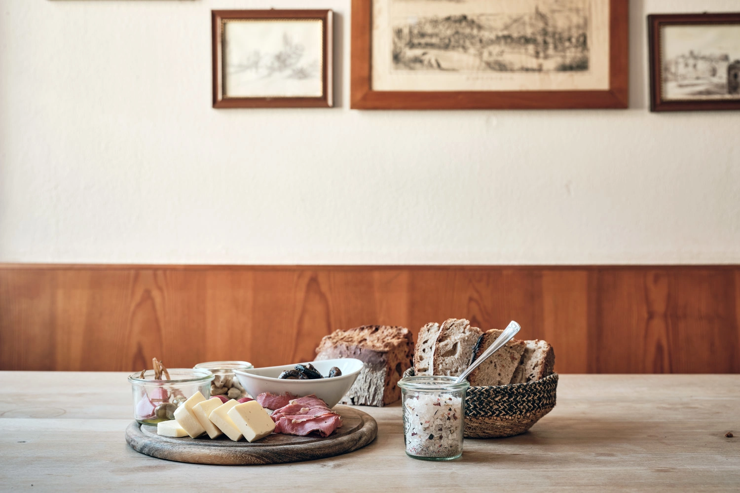 Auf einem Tisch ist eine Holzplatte mit Käse und Wurst sowie weiteren Snacks angerichtet. Daneben steht ein Korb mit Brotscheiben.
