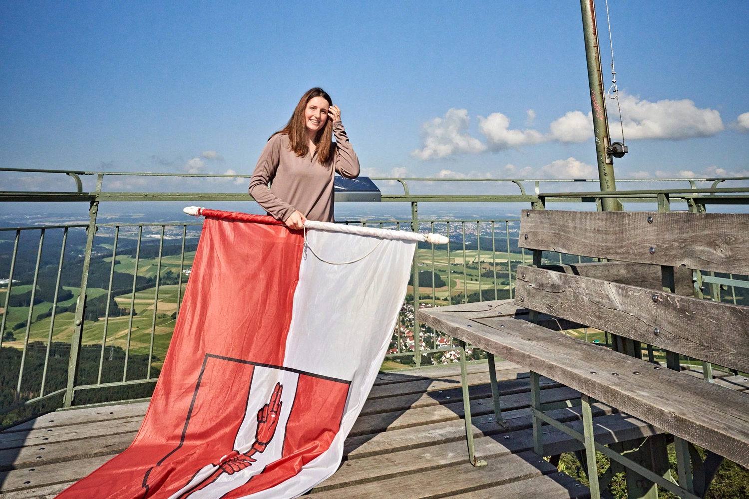 Person steht auf der Aussichtsplattform des Lembergturms und hält eine große rot-weiße Fahne, im Hintergrund die weite Landschaft der Schwäbischen Alb unter blauem Himmel.
