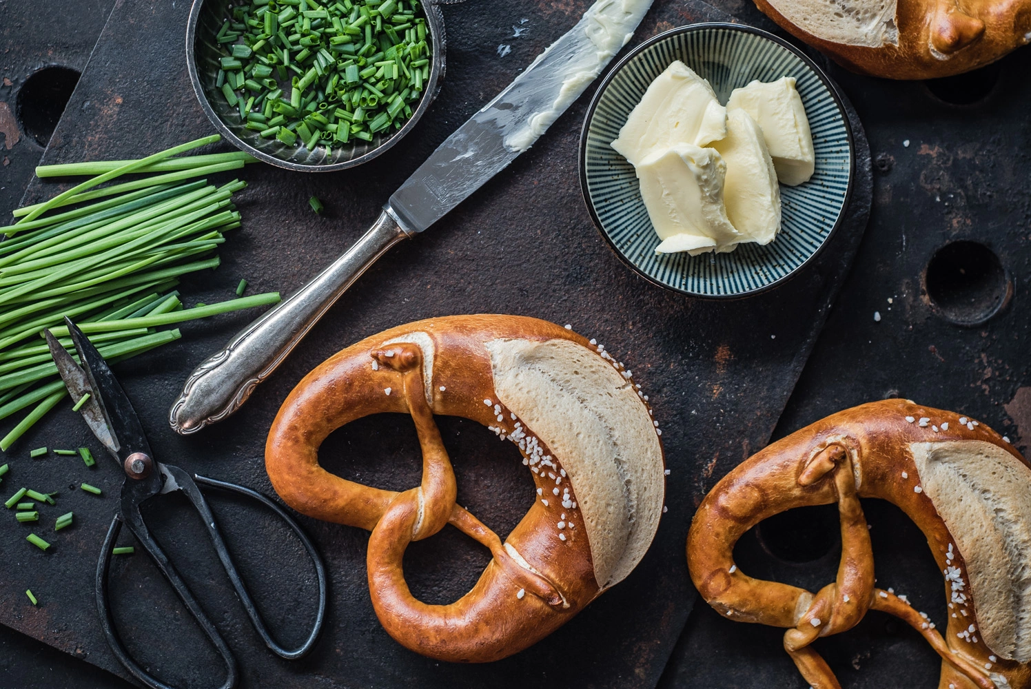 Frische Brezeln mit grobem Salz, dazu Butterstückchen auf einem Teller, ein Messer mit Butterspuren und gehackter Schnittlauch auf dunkler Unterlage – typisch schwäbischer Genuss.