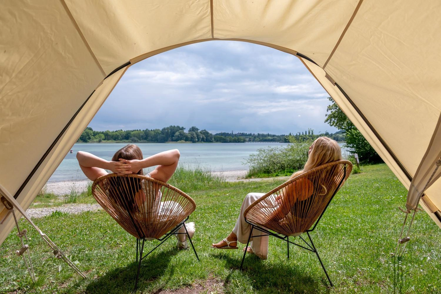 Blick aus einem geöffneten Zelt auf zwei Personen in Lounge-Stühlen am Bodenseeufer mit grüner Wiese und ruhigem Wasser unter leicht bewölktem Himmel.