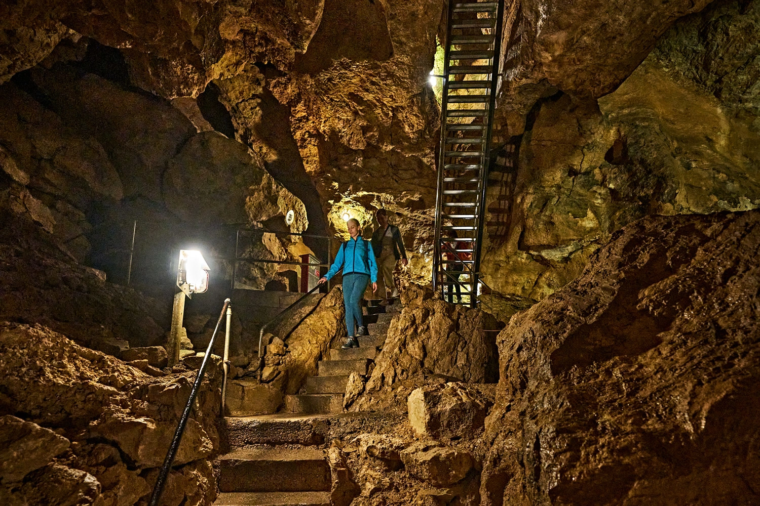 Person steigt über beleuchtete Steintreppen und Metallleitern durch die weitläufige, felsige Tiefenhöhle der Schwäbischen Alb.