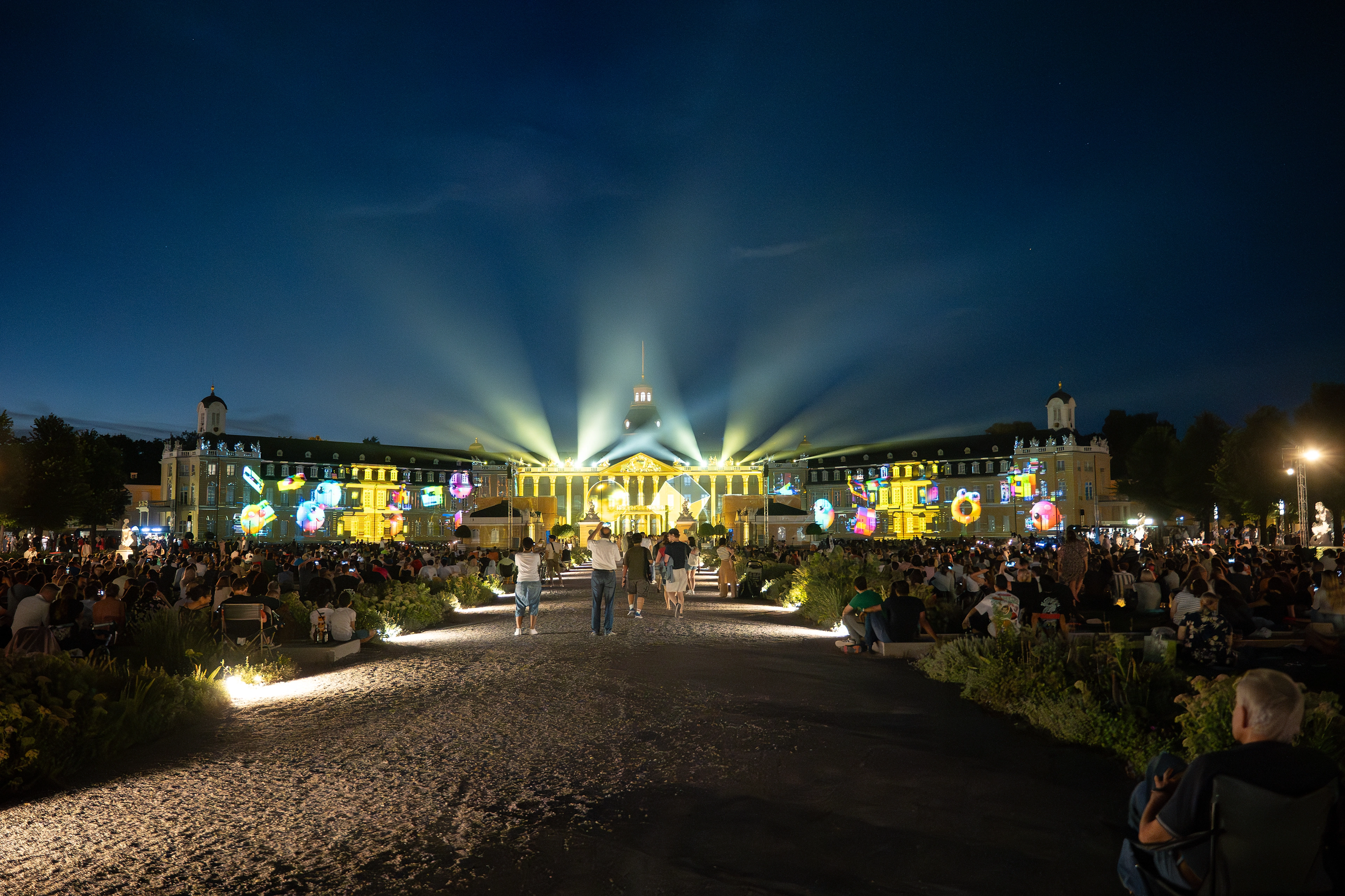 Menschenmenge im Schlossgarten betrachtet die bunten Videoprojektionen der Schlosslichtspiele auf der Fassade des Karlsruher Schlosses bei Nacht.