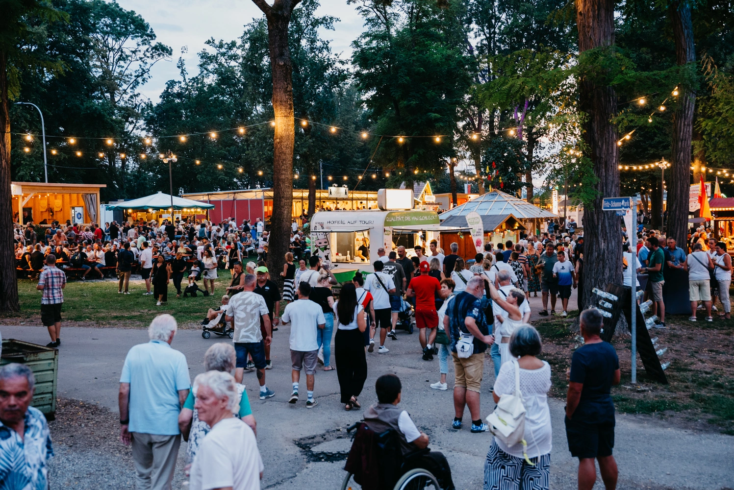 Besucher auf einem Festgelände in einem Park mit vielen Bäumen