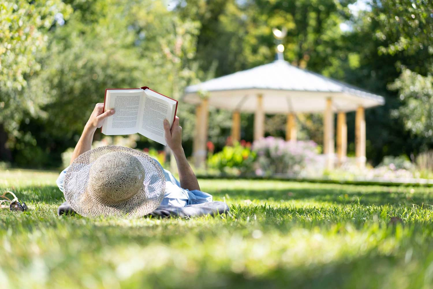 A woman lies relaxed on a meadow and reads a book in the shade of the trees. 