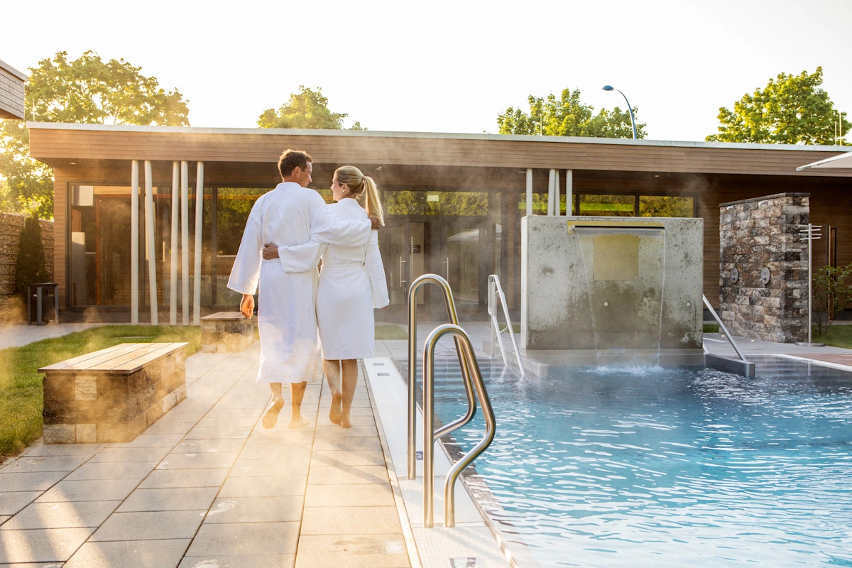 A couple walks arm in arm in their bathrobes along an outdoor pool.