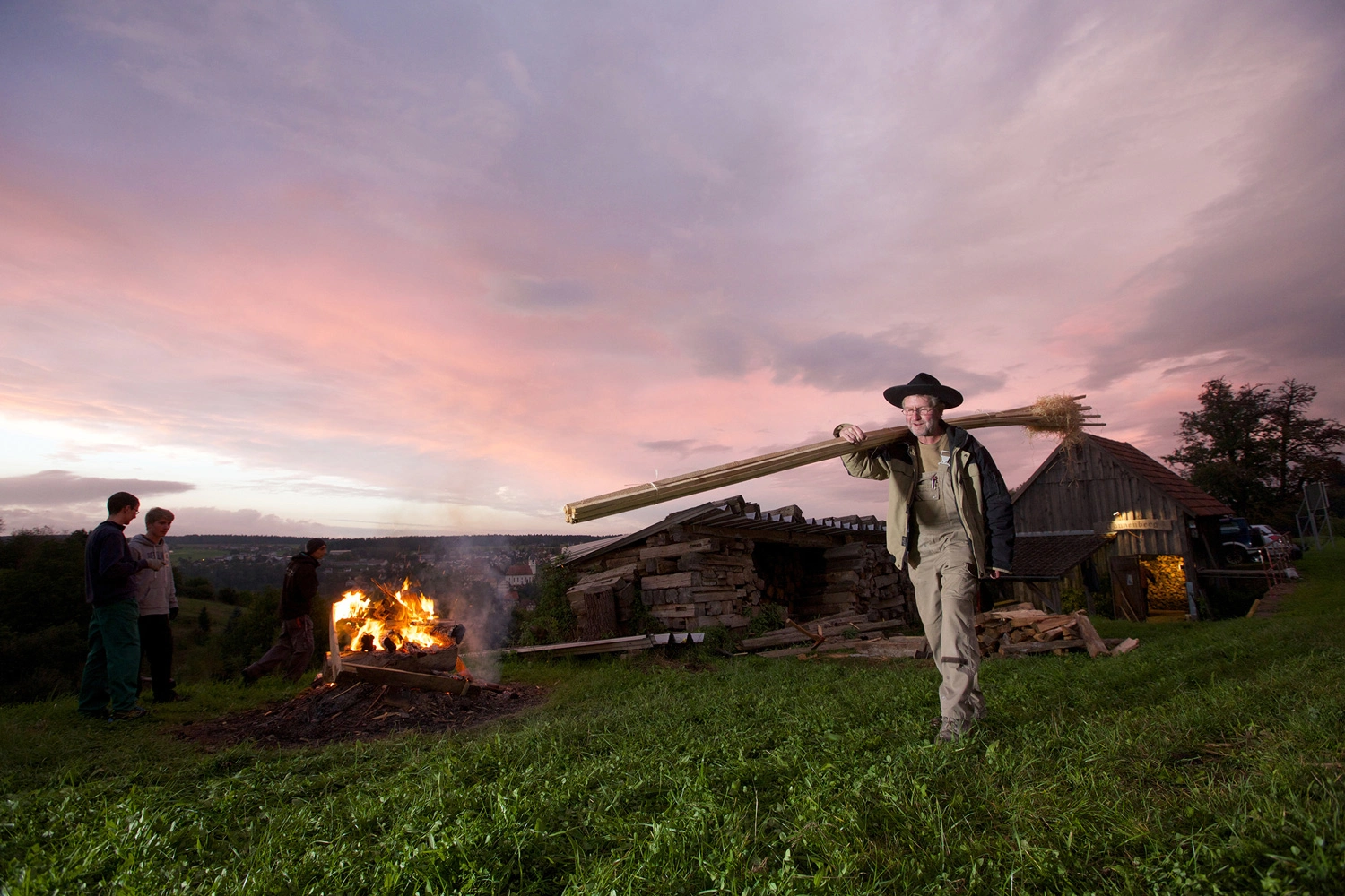 A man carries wooden planks with straw past a campfire.