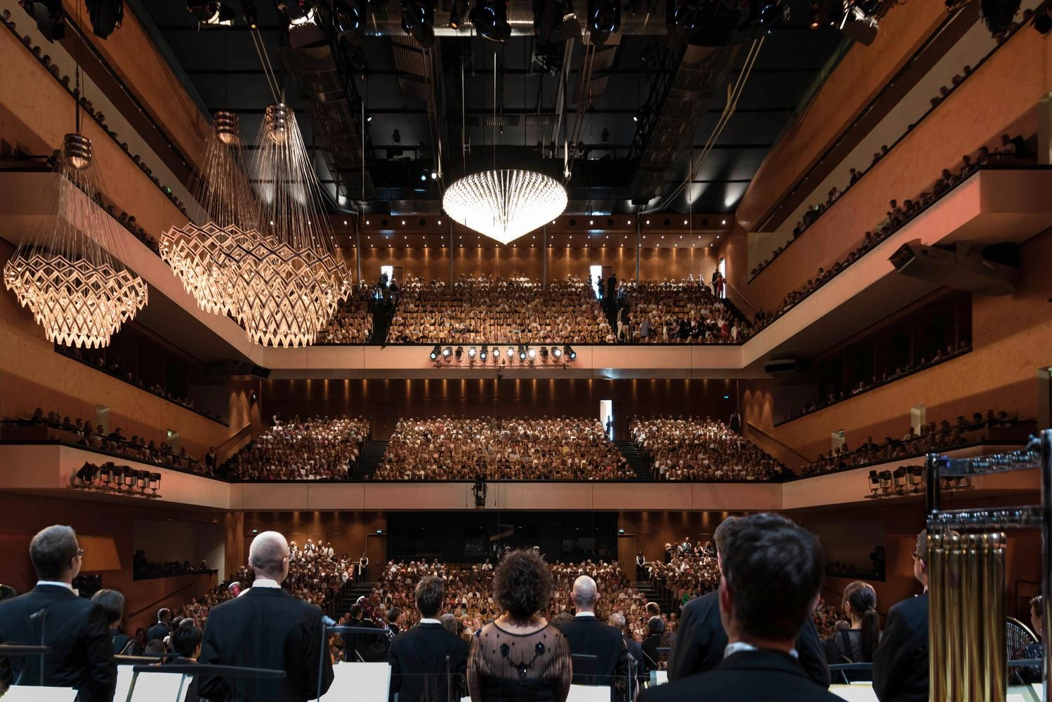 View from the stage into a large, modern concert hall with several tiers, bright chandeliers, and fully occupied seats; musicians are standing in the foreground.
