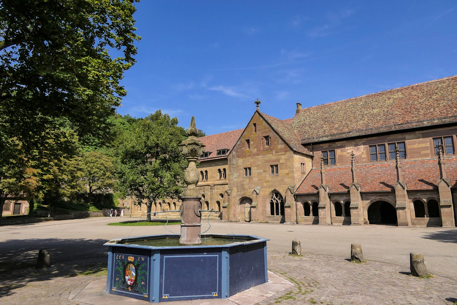 Historic courtyard of Maulbronn Monastery with an ornate fountain and medieval sandstone buildings beneath a blue sky.