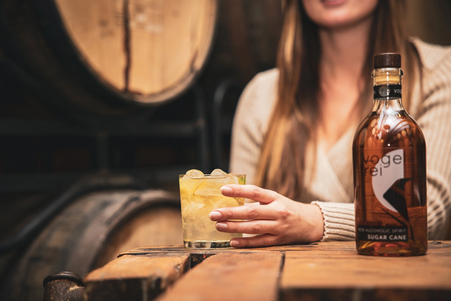A bottle of non-alcoholic rum labeled Heimat Vogelfrei Sugar Cane stands on a table. A woman in the background holds a glass of rum in her left hand.