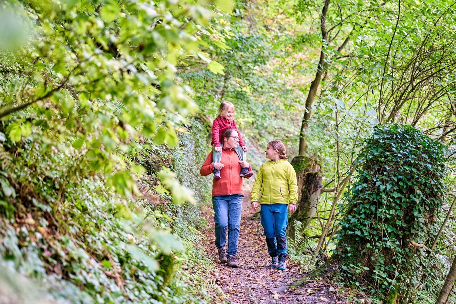 Three people are walking together on a narrow forest path, surrounded by dense greenery and autumn leaves.