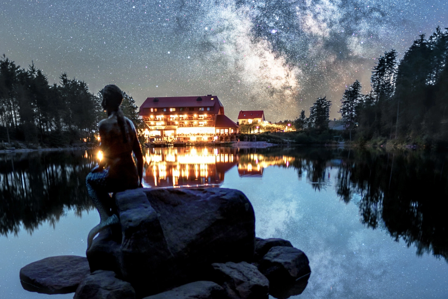 Night scene at the lake with illuminated building, clear reflection in the water, and a sculpture in the foreground against a sparkling starry sky.