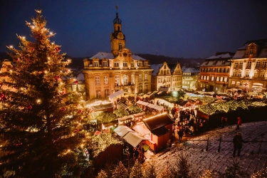 View of a lively Christmas market at dusk on the historic market square in Schwäbisch Hall. The square is surrounded by traditional buildings and illuminated by warm lights.