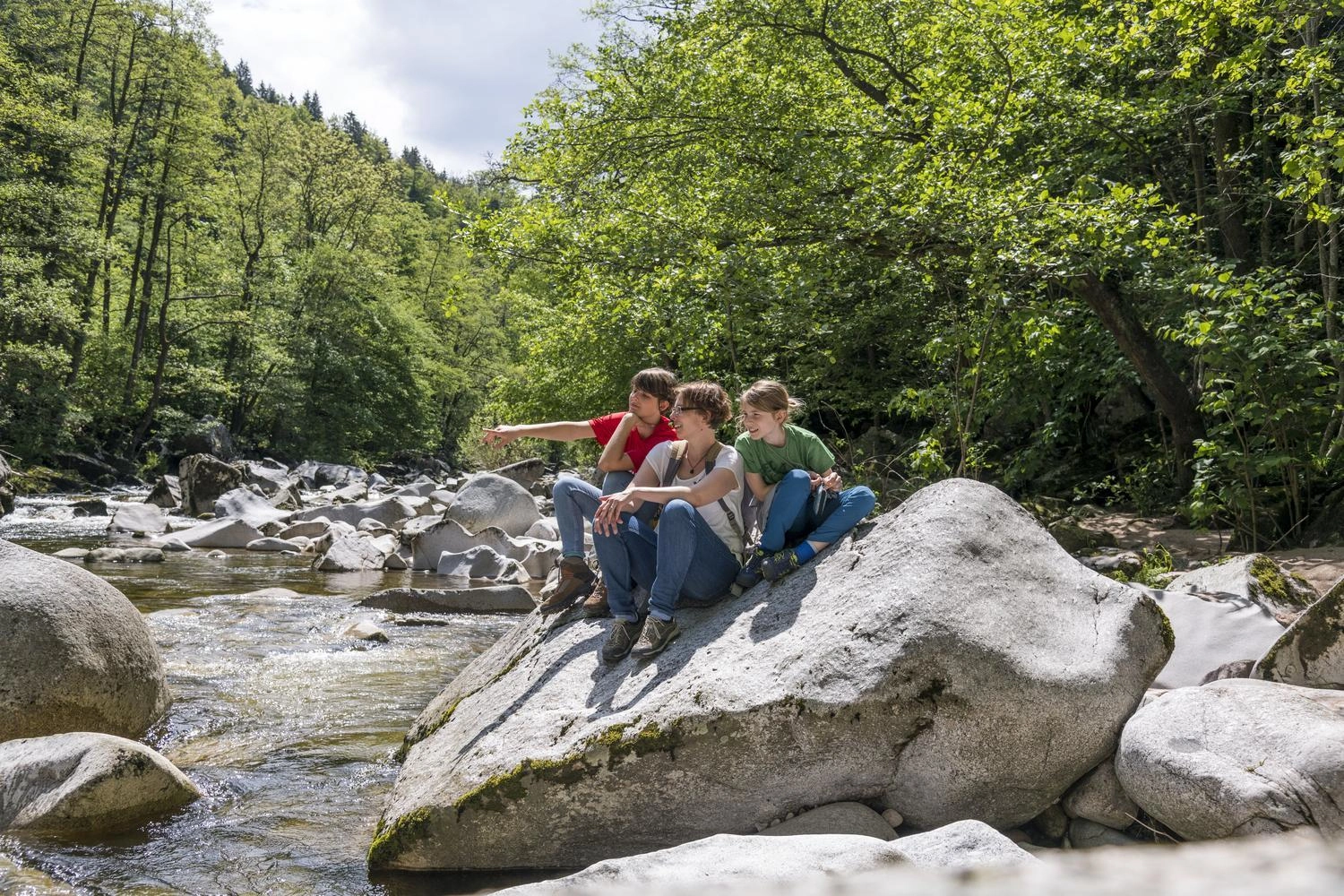 A mother and her two children are sitting on a large stone in a riverbed.