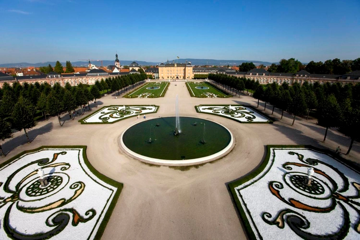 Luftaufnahme einer prunkvollen Gartenanlage mit Wasserspielen. Am Ende der Anlage ist das Schloss zu erkennen.