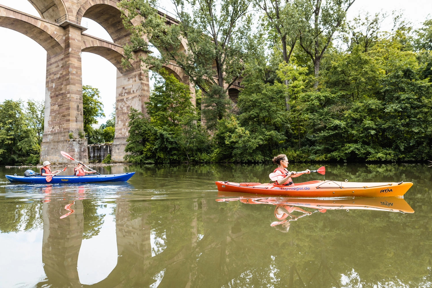 Eine Frau und zwei Kinder fahren in zwei Kanus auf einem Fluss unter einem Viadukt hindurch.