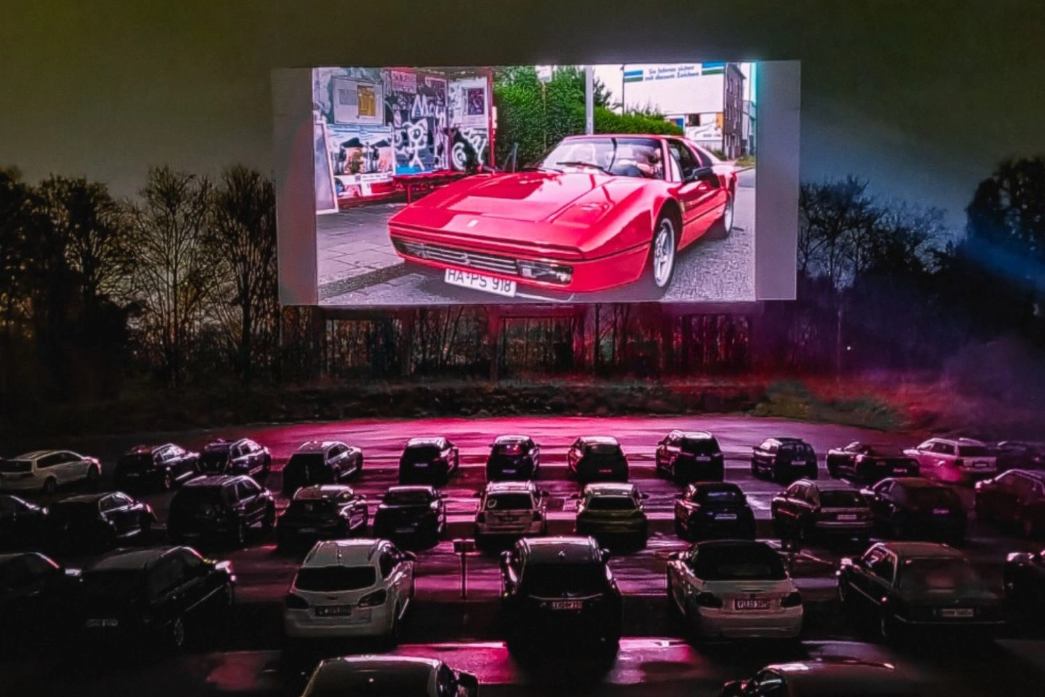 A large car cinema screen is shown at night. Rows of cars are parked in front of it.