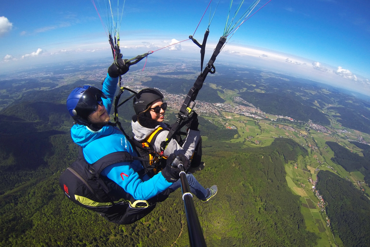 Two people glide high above a green mountain landscape in tandem paragliding, with sweeping views over valleys and villages.