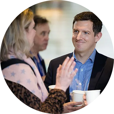 Group of employees conversing indoors at an event, holding paper cups and engaging in discussion with others in the background.