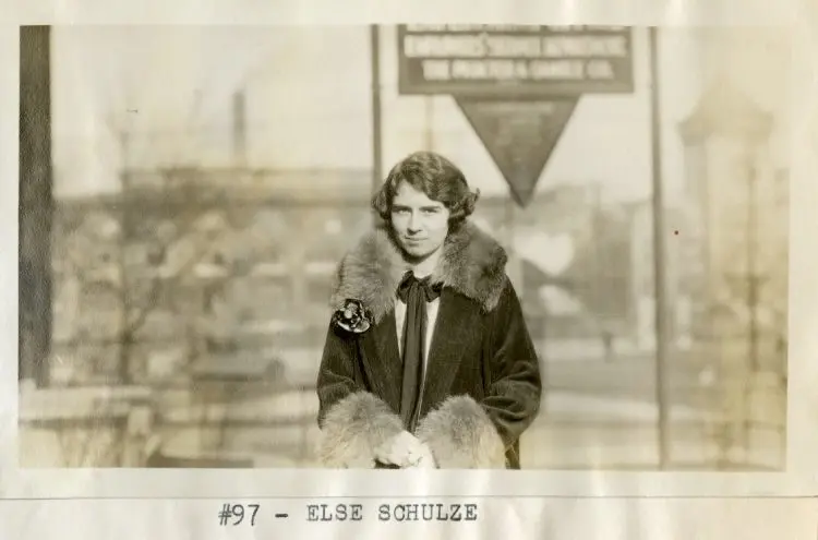A woman with short dark hair in a coat. She holds her hands together and stands outdoors with a blurred urban background and a triangular sign behind her.