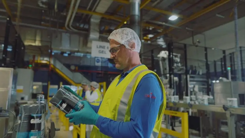 Photograph shows a factory worker wearing safety glasses, hairnet, gloves, and high-visibility vest while inspecting a container. Industrial production line, machinery, overhead pipes, and another worker provide manufacturing context.