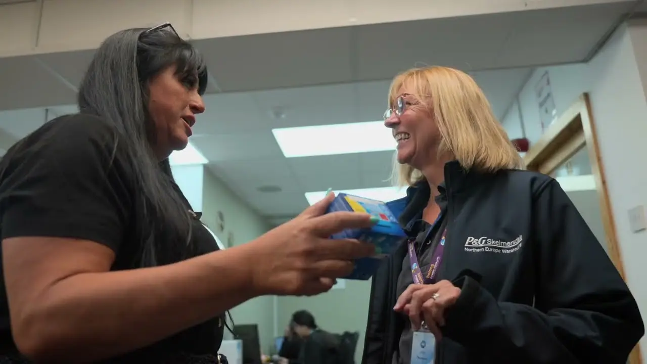 Two women smiling and talking in a hallway, with one handing a packaged product to a P&G employee wearing a company jacket.