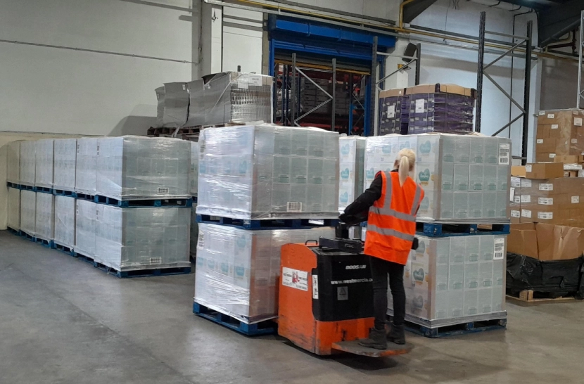 Warehouse scene with worker in high-visibility vest operating electric pallet jack among stacked pallets, ensuring efficient logistics management for brand products.