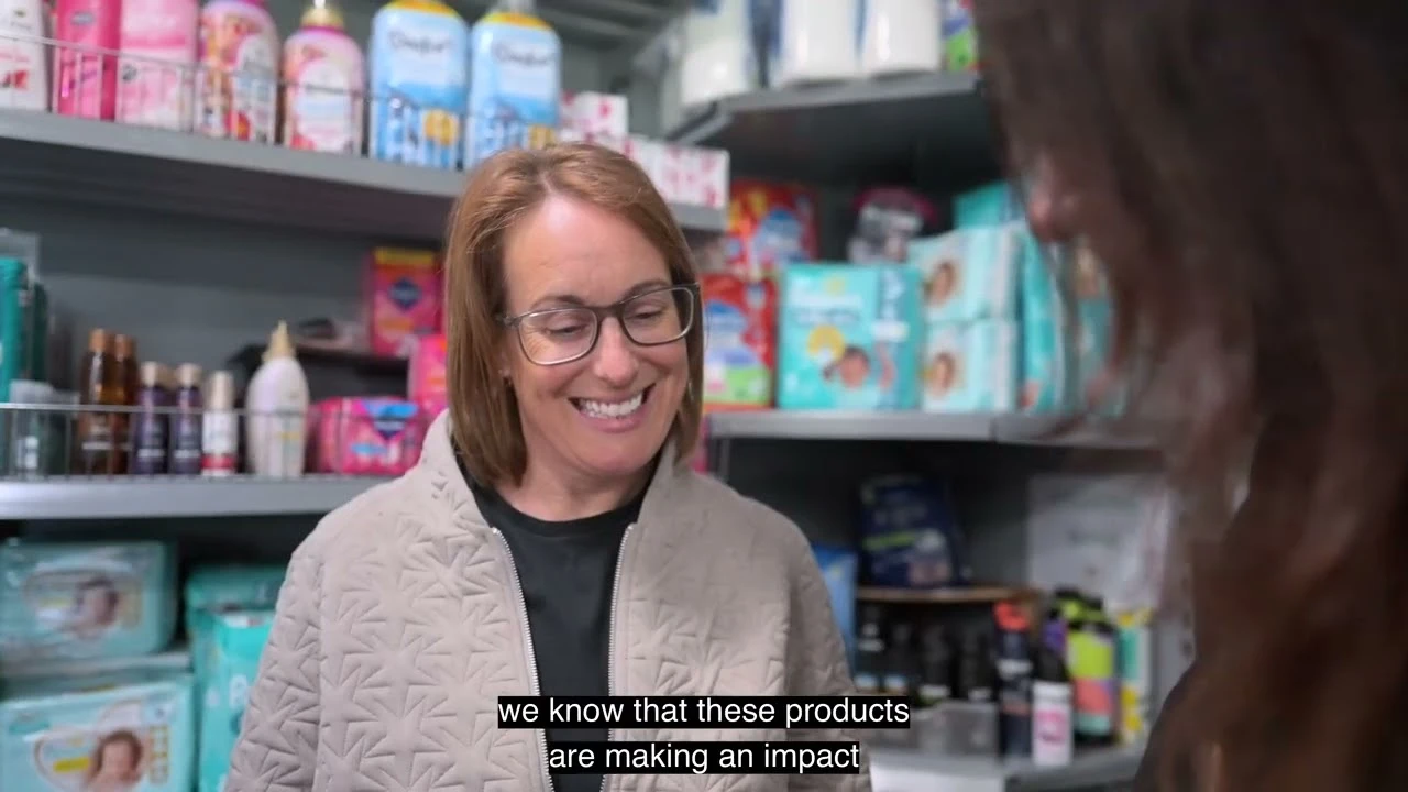 Smiling woman with glasses standing in a retail store aisle stocked with personal care and baby products.