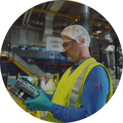 Photograph shows a factory worker wearing safety glasses, hairnet, gloves, and high-visibility vest while inspecting a container. Industrial production line, machinery, overhead pipes, and another worker provide manufacturing context.