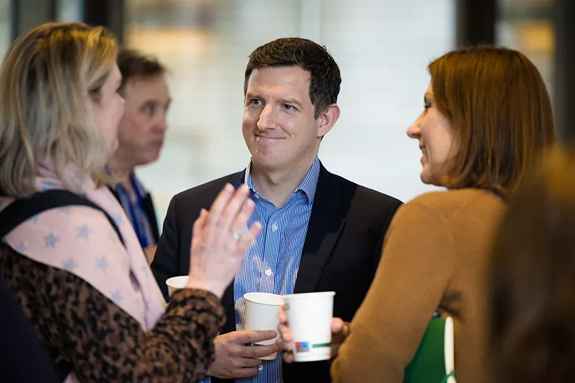 Group of employees conversing indoors at an event, holding paper cups and engaging in discussion with others in the background.