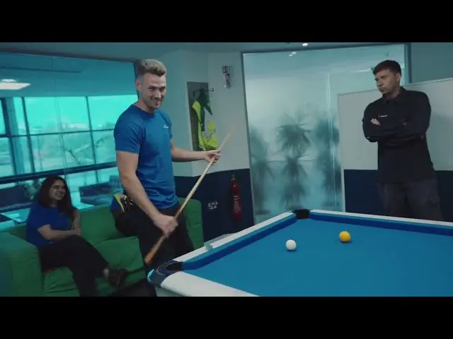 Photograph shows a person holding a pool cue beside a billiards table in a modern office lounge area, while two other people observe nearby. Glass walls, office seating, and large windows are visible in the background.