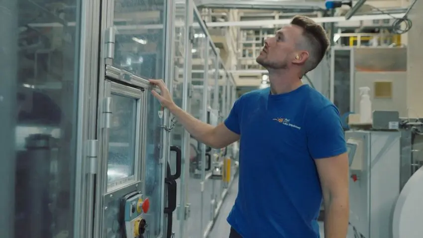 Photograph showing a factory worker inspecting enclosed industrial machinery inside a manufacturing facility. Worker wears a blue shirt and safety earplug beside glass panels, control buttons, warning indicators, pipes, and production equipment.