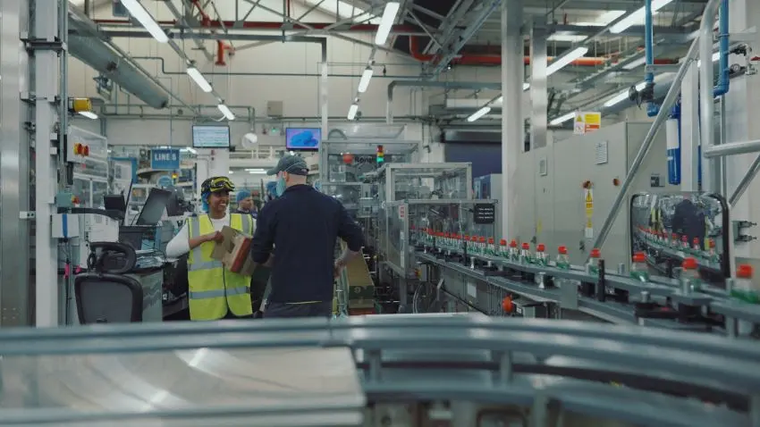 Photograph shows a modern manufacturing facility with two workers beside automated machinery and conveyor lines. Safety vests, control panels, and equipment highlight an active production environment.