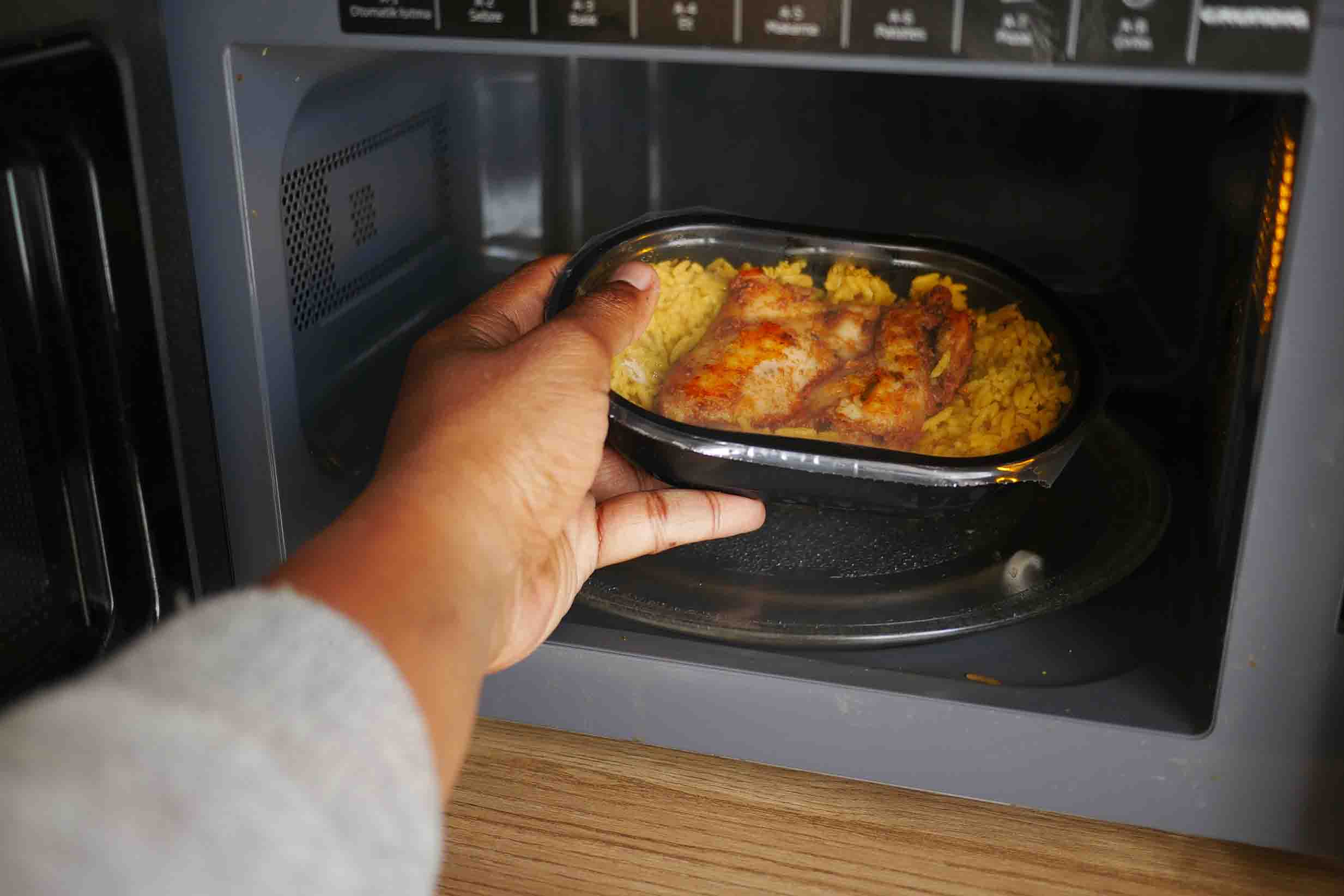 A hand holding a prepared meal in a plastic bowl reaching into a microwave oven.