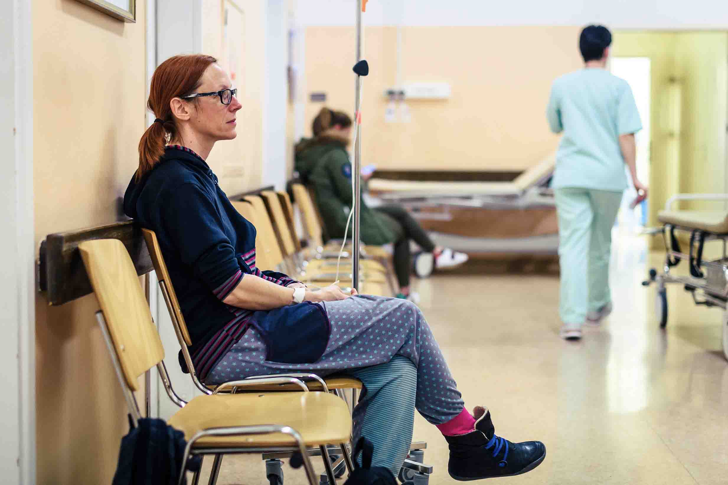 A woman with red hair wearing a navy-blue sweatshirt and blue pants, sits in a hospital waiting room, looking into the distance. 