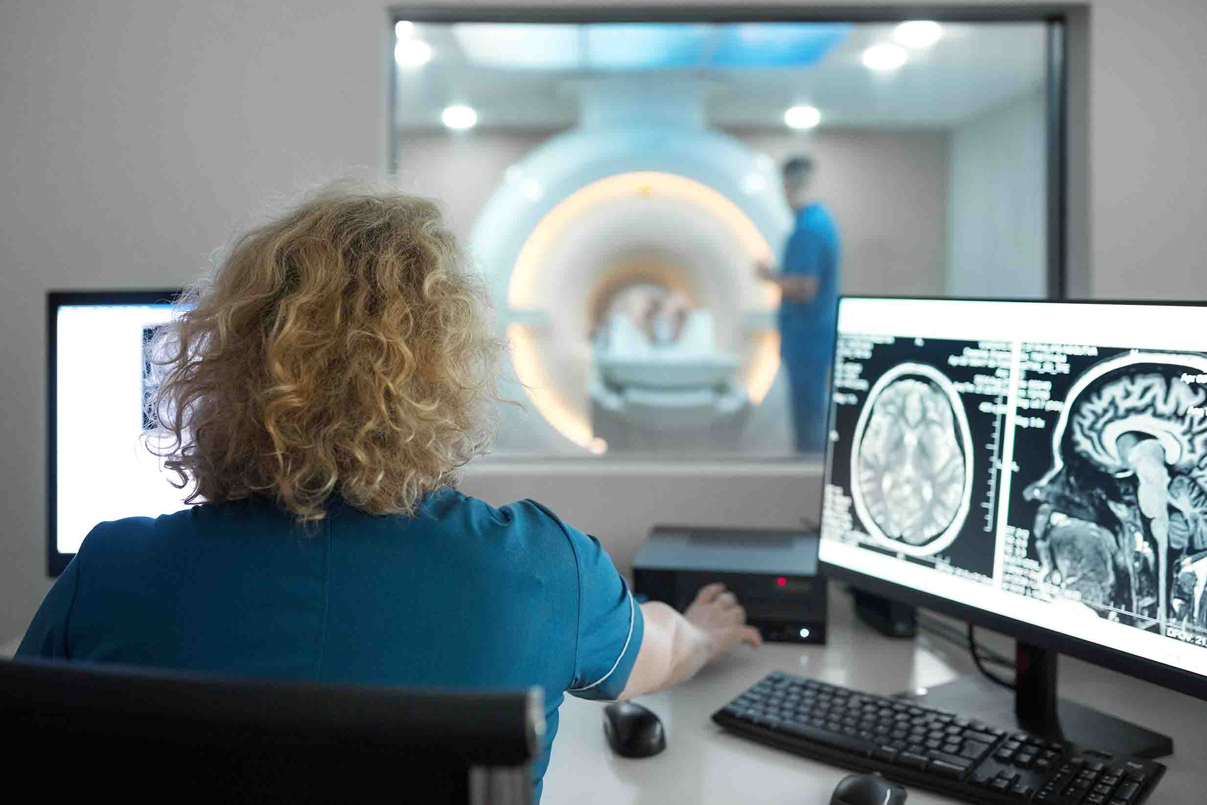 A woman with blonde hair wearing blue scrubs looking at images of a brain MRI.