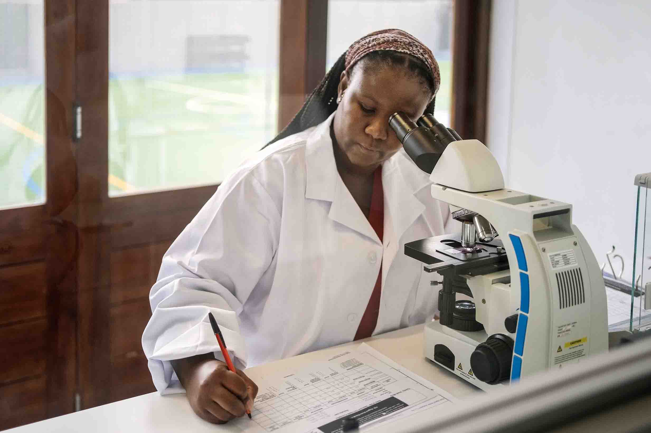 A Black woman scientist taking notes at a microscope.