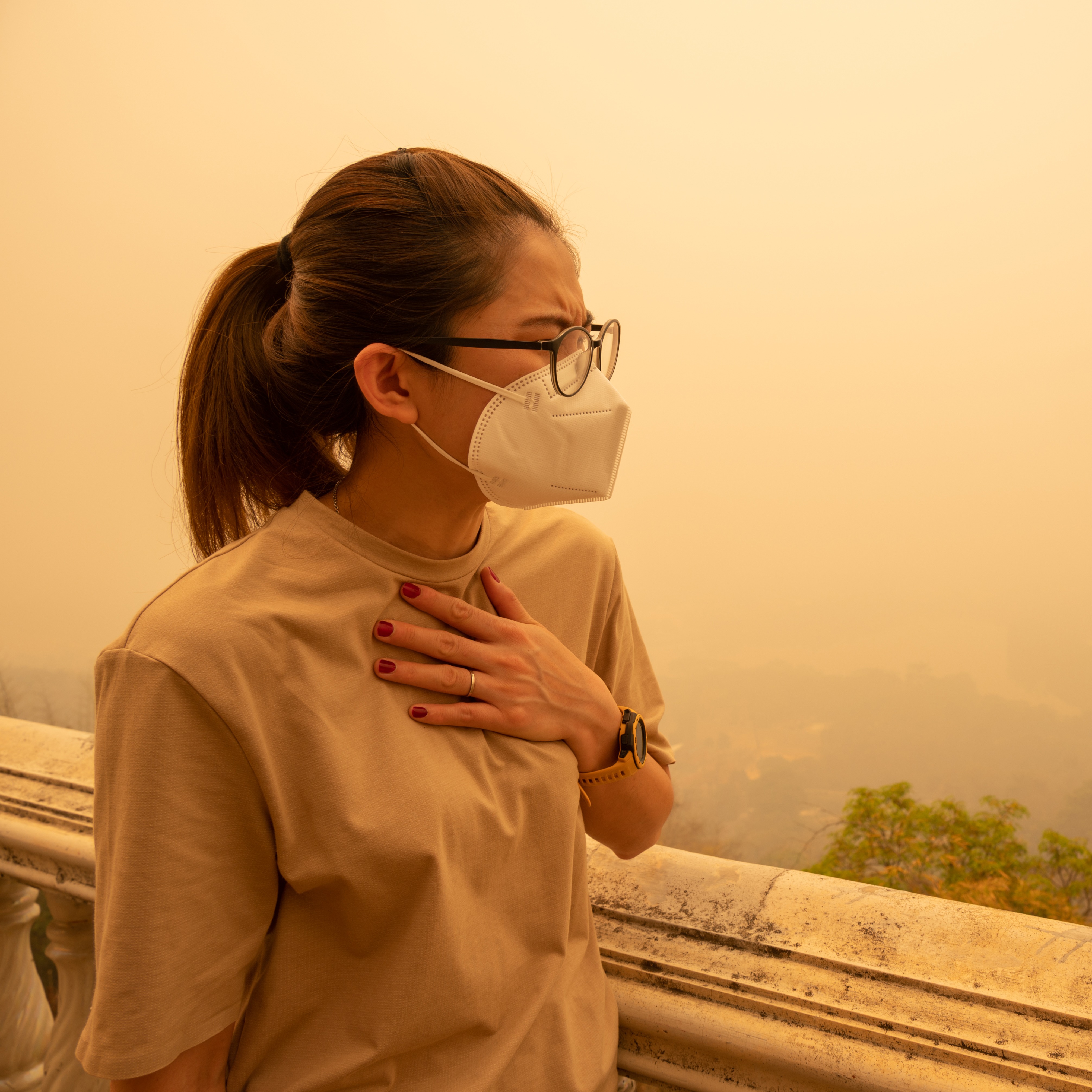 Person in mask stands with one hand on chest, looking at hazy sky.