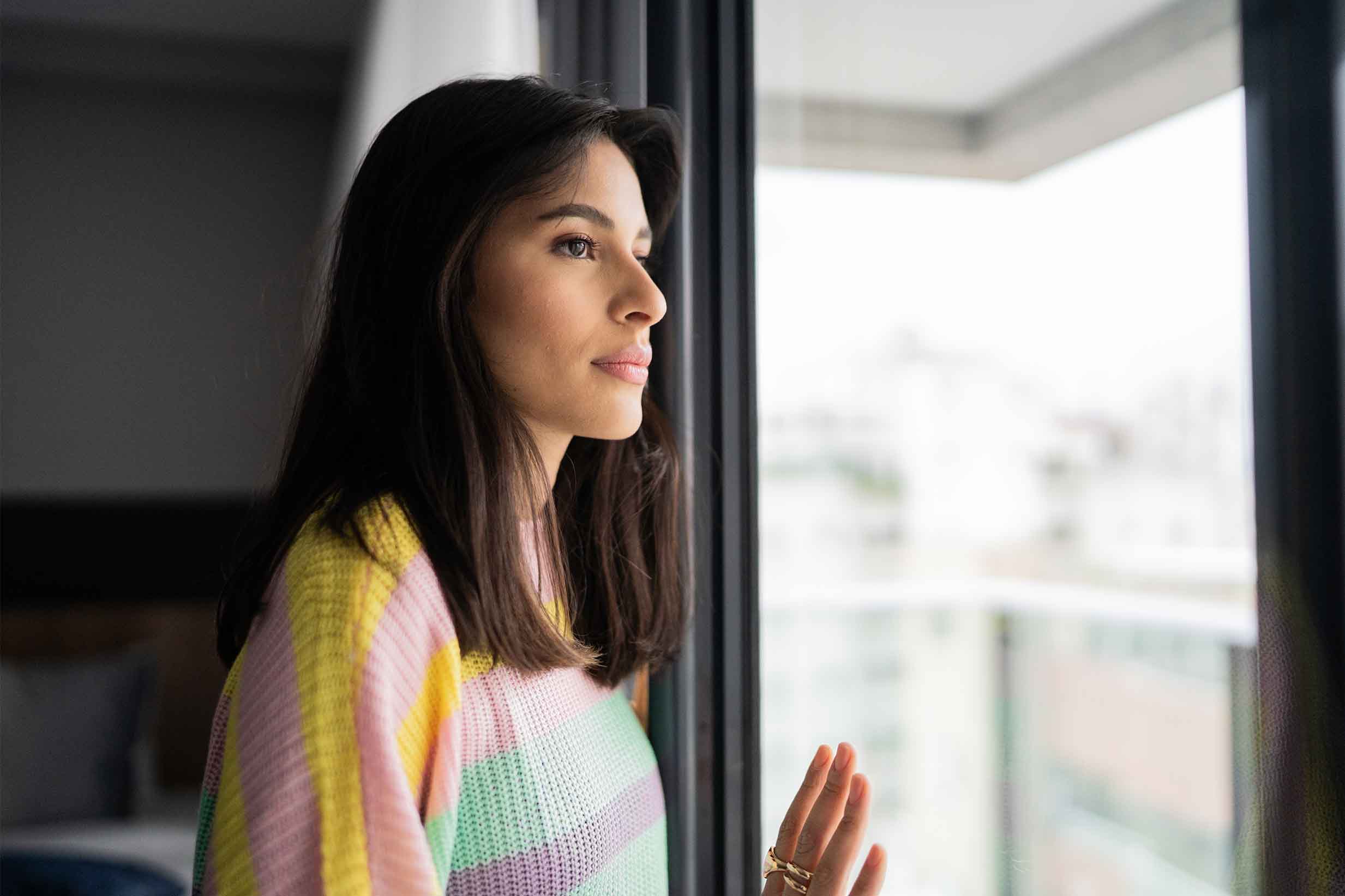 A woman with shoulder-length brown hair stands at a large window, looking out with a solemn expression